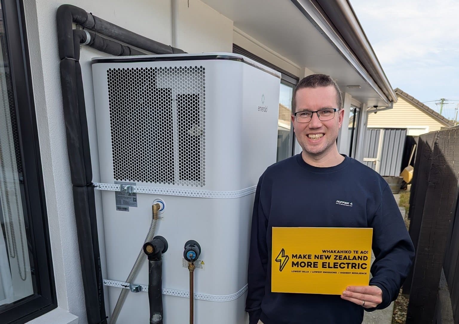 A smiling homeowner beside their new hot water heat pump, holding a Make New Zealand More Electric sign